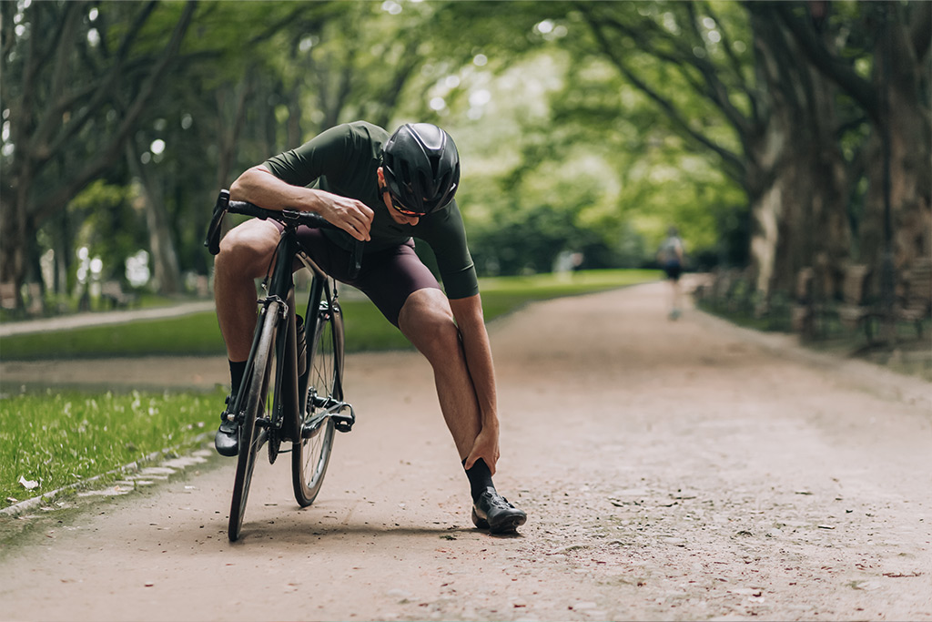 Cyclist in a park stopping to hold his ankle, showing a fall sports injury and muscle discomfort, seeking relief with Massage in Maple Ridge